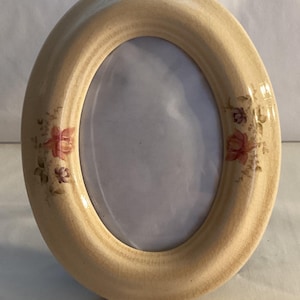 May include: Oval-shaped ceramic picture frame in a cream color with a glossy finish. The frame is decorated with small pink and purple floral accents. The frame is designed to hold an oval-shaped photograph.