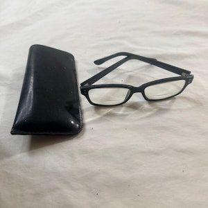 May include: Black reading glasses with a rectangular frame and clear lenses, next to a black leather glasses case. The glasses have a classic design and are suitable for reading. The case is rectangular and has a smooth, shiny surface.
