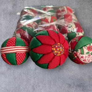 May include: Handmade Christmas baubles in red and green. The baubles feature patterns like polka dots, stripes, and floral prints. One bauble has a poinsettia design. A clear bag holds more baubles, creating a festive holiday display.