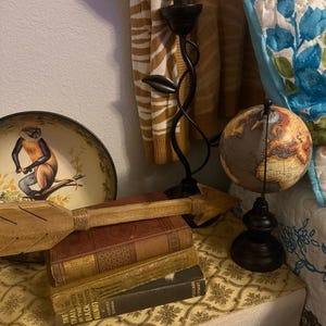 May include: A decorative arrangement featuring a small globe, a wooden arrow, and a stack of vintage books. A black metal candle holder with a leaf design and a decorative plate with a monkey illustration are also present. The items rest on a patterned surface.