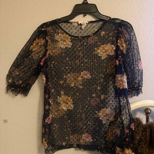 May include: A sheer black blouse with a floral pattern in shades of yellow, pink, and brown. The blouse has short sleeves and a round neckline. The fabric has a dotted texture.