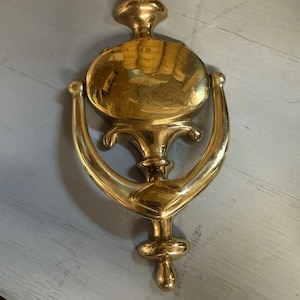 May include: A polished brass door knocker featuring a vertical, elongated design with a central oval striking plate and a curved, U-shaped handle. The metal surface is reflective and smooth, showcasing a metallic gold tone throughout the piece.
