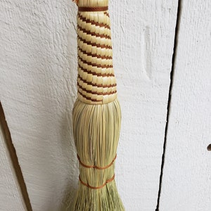 May include: A handmade broom with a natural bristle head and a woven handle with brown accents.
