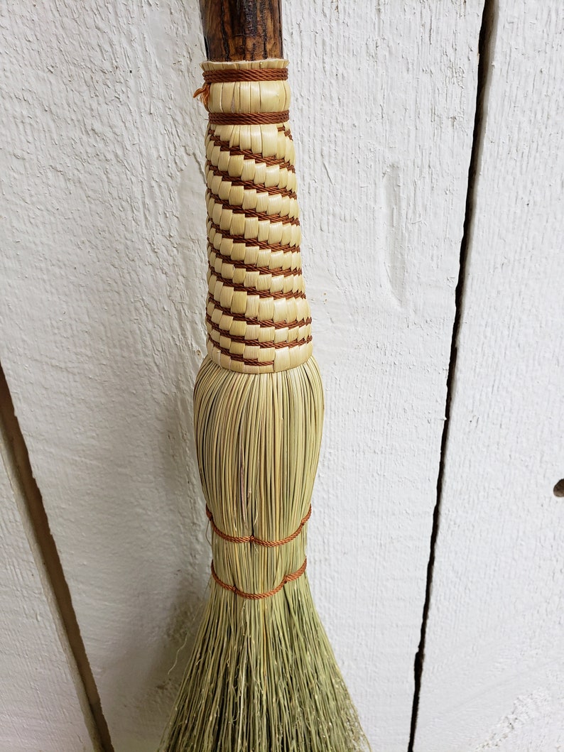 May include: A handmade broom with a natural bristle head and a woven handle with brown accents.