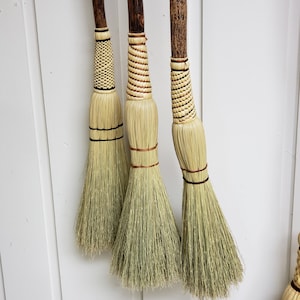 May include: Three handmade brooms with natural straw bristles and wooden handles. The handles are wrapped with a decorative pattern of brown and white twine.