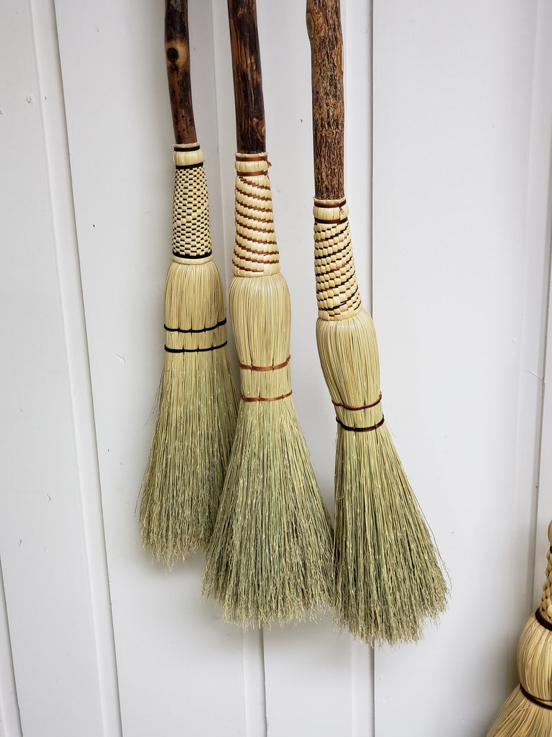 May include: Three handmade brooms with natural straw bristles and wooden handles. The handles are wrapped with a decorative pattern of brown and white twine.