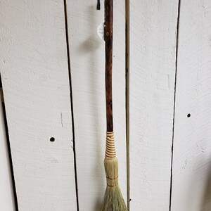 May include: A brown wooden-handled broom with a natural fiber brush. The broom is hanging on a white wooden wall.