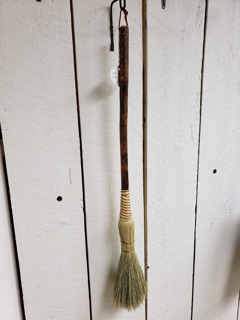 May include: A brown wooden-handled broom with a natural fiber brush. The broom is hanging on a white wooden wall.