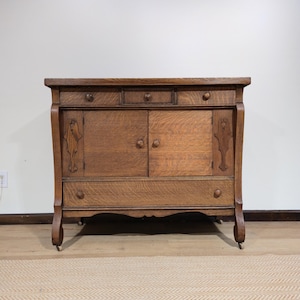 May include: A brown wooden sideboard featuring three top drawers, two central cabinet doors with round knobs, and one large bottom drawer. The piece has curved side supports, decorative vertical carvings, and small wheels at the base of each leg.