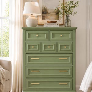 May include: A sage green chest of drawers with gold-toned hardware. The tall, wooden dresser has seven drawers of varying sizes. A white lamp and framed artwork sit on top of the dresser. The chest is a stylish storage solution for a bedroom.