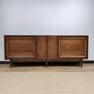 May include: A long, rectangular wooden sideboard featuring two outer cabinets with framed panels and a central section with two doors displaying a diamond-shaped wood grain pattern. The piece is supported by four tapered legs and includes the text www.ed-furnituredesign.com.