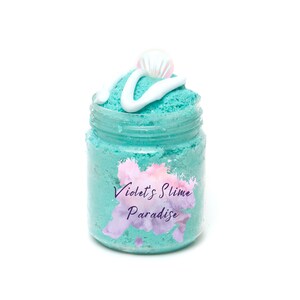 May include: A teal blue slime in a clear glass jar with a white swirl on top and a white seashell. The jar has a label that says "Violet's Slime Paradise".
