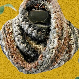 May include: A handmade, chunky knit cowl in neutral tones of gray, brown, and white. The infinity scarf is displayed on a black hanger against a yellow background. The soft, textured yarn creates a warm and cozy accessory.