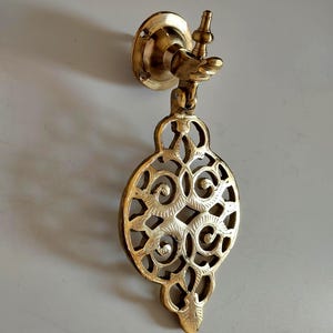 May include: Ornate brass door knocker with a decorative, openwork design. The knocker features a circular shape with intricate swirling patterns and a small handle. The brass has a warm, golden hue.