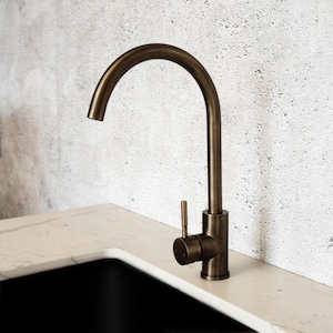 May include: A bronze kitchen tap with a curved spout and a single lever handle. The tap is mounted on a light-coloured countertop, with a black sink visible below. The background is a textured, light grey wall.