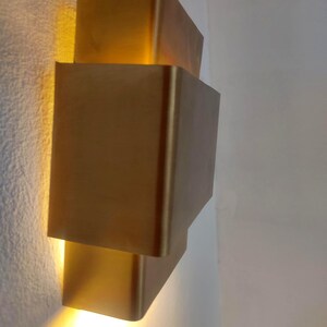May include: A modern, gold-colored wall sconce with a stacked, geometric design. The sconce emits a warm, yellow light, creating a soft glow. The fixture is mounted on a white wall, adding a touch of contemporary style to any space.