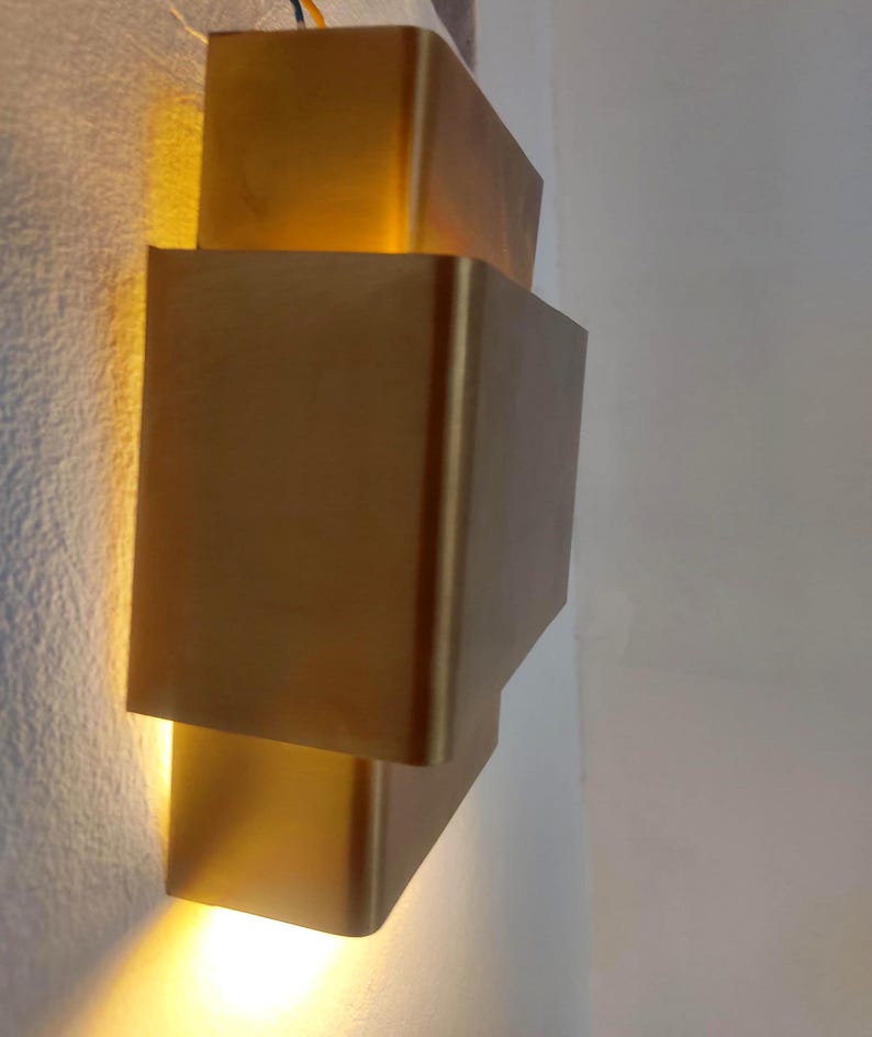 May include: A modern, gold-colored wall sconce with a stacked, geometric design. The sconce emits a warm, yellow light, creating a soft glow. The fixture is mounted on a white wall, adding a touch of contemporary style to any space.