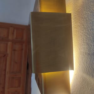 May include: A modern, gold-toned wall sconce with a rectangular design. The sconce is mounted on a textured, off-white wall and emits a warm, inviting light. The fixture has a layered appearance, creating depth and visual interest.