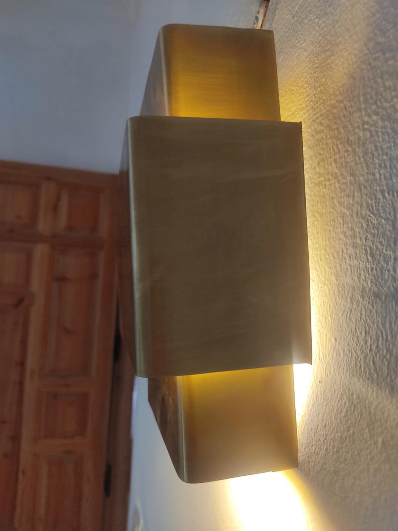 May include: A modern, gold-toned wall sconce with a rectangular design. The sconce is mounted on a textured, off-white wall and emits a warm, inviting light. The fixture has a layered appearance, creating depth and visual interest.