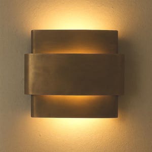 May include: A bronze-colored wall sconce with a layered design. The sconce has a rectangular shape with three stacked sections, emitting a warm, inviting light. The fixture is mounted on a neutral-toned wall, creating a modern aesthetic.