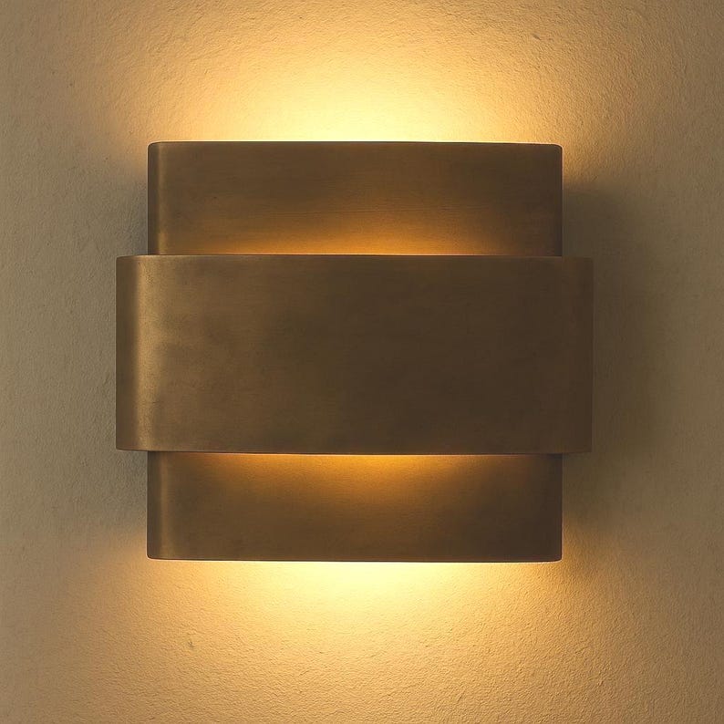 May include: A bronze-colored wall sconce with a layered design. The sconce has a rectangular shape with three stacked sections, emitting a warm, inviting light. The fixture is mounted on a neutral-toned wall, creating a modern aesthetic.