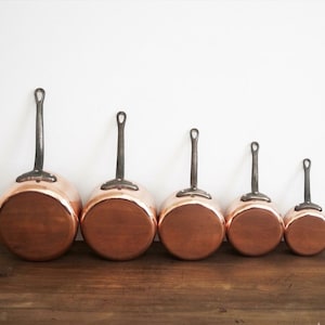 May include: A set of five copper saucepans with long, dark metal handles. The pans are arranged in a row, decreasing in size from left to right. The copper has a shiny, reflective surface, and the handles have a loop at the end.