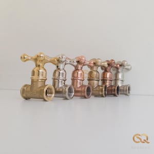 May include: A row of six metal water faucets with cross-shaped handles, displayed in a gradient of finishes including polished brass, chrome, copper, antique bronze, rose gold, and brushed nickel, each featuring a threaded 1/2 inch pipe connection.