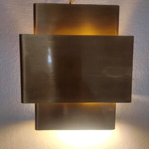 May include: A rectangular, brass-toned wall sconce with a layered design. The sconce has three horizontal sections, with the middle section slightly protruding. The light source is visible at the top and bottom, creating a warm glow.