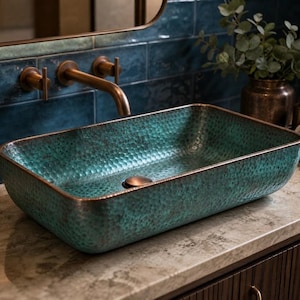 Op de afbeelding: Een rechthoekige waskom met een gehamerde textuur en een verweerde groenblauwe en koperen patina afwerking. De waskom heeft afgeronde hoeken en een centrale ronde afvoerplug in een bijpassende koperkleur, geplaatst tegen een blauwe tegelwand.