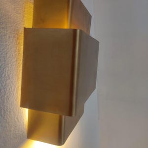 May include: A modern, gold-colored wall sconce with a stacked, rectangular design. The sconce emits a warm, yellow light, creating a soft glow. The fixture is mounted on a white wall.