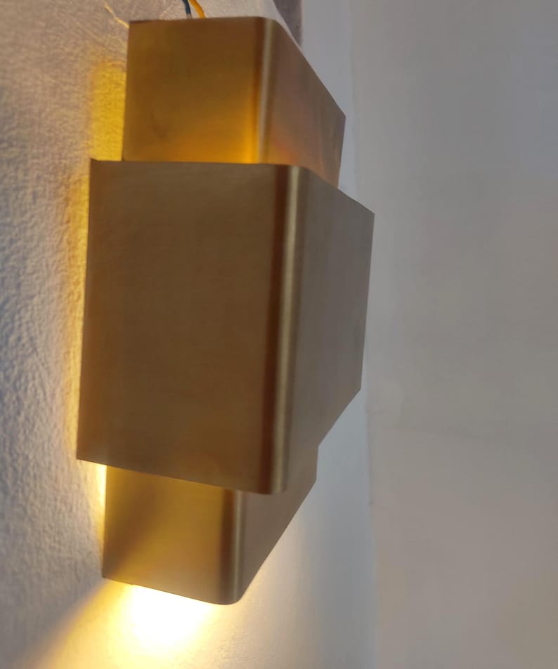 May include: A modern, gold-colored wall sconce with a stacked, rectangular design. The sconce emits a warm, yellow light, creating a soft glow. The fixture is mounted on a white wall.