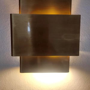 May include: A modern wall sconce with a rectangular design. The sconce is made of brushed brass and emits a warm, inviting light. The fixture has a layered design, with a larger rectangular shade over a smaller one.