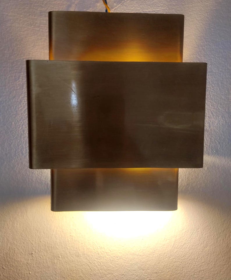 May include: A modern wall sconce with a rectangular design. The sconce is made of brushed brass and emits a warm, inviting light. The fixture has a layered design, with a larger rectangular shade over a smaller one.