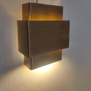 May include: A modern, geometric wall sconce with a warm, golden glow. The fixture is composed of stacked, rectangular blocks in a metallic finish. The light shines from the spaces between the blocks, creating a layered effect. The sconce is mounted on a white wall.
