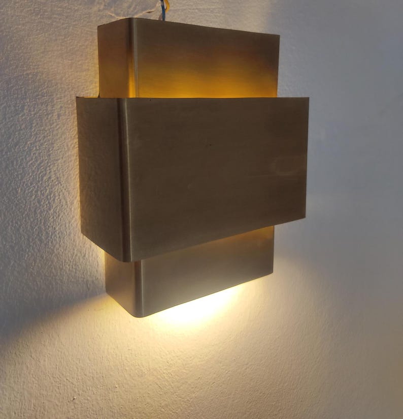 May include: A modern, geometric wall sconce with a warm, golden glow. The fixture is composed of stacked, rectangular blocks in a metallic finish. The light shines from the spaces between the blocks, creating a layered effect. The sconce is mounted on a white wall.