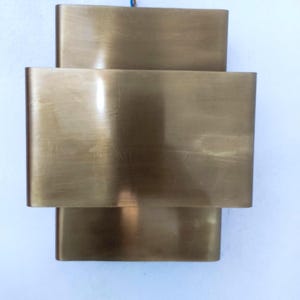 May include: A brass-colored wall sconce with a layered rectangular design. The sconce has three stacked, curved rectangular sections, creating a modern, geometric aesthetic. The metal has a brushed finish, reflecting light. The fixture is mounted on a white surface.