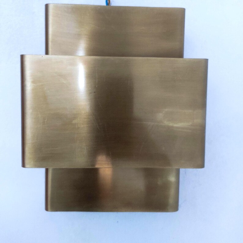 May include: A brass-colored wall sconce with a layered rectangular design. The sconce has three stacked, curved rectangular sections, creating a modern, geometric aesthetic. The metal has a brushed finish, reflecting light. The fixture is mounted on a white surface.