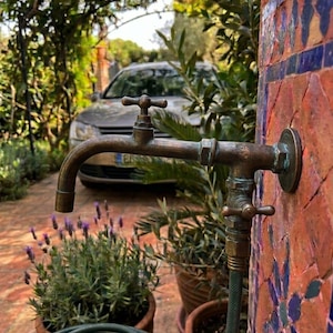 May include: A weathered bronze outdoor tap mounted on a mosaic tile wall. The tap features a cross-handle top and a secondary lower valve connected to a green garden hose. The metal has a dark, aged patina with visible signs of oxidation.