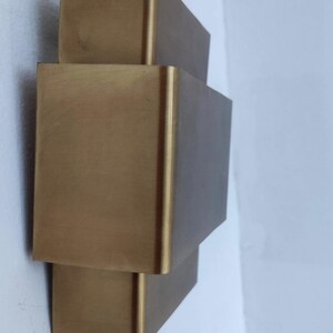 May include: A gold-colored, geometric wall sconce with a stacked rectangular design. The fixture is mounted on a white wall, and wires are visible at the top. The sconce has a modern, minimalist aesthetic.