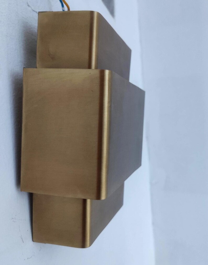 May include: A gold-colored, geometric wall sconce with a stacked rectangular design. The fixture is mounted on a white wall, and wires are visible at the top. The sconce has a modern, minimalist aesthetic.