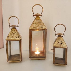 Puede incluir: Tres faroles de metal dorado de diferentes tamaños. Cada farol tiene un asa, una ventana de cristal y patrones decorativos recortados. Una vela encendida está dentro del farol central. Los faroles son probablemente para fines decorativos.