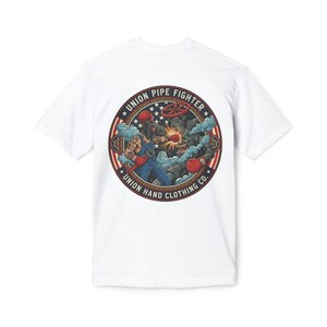 Könnte beinhalten: Weißes T-Shirt mit einem kreisförmigen Grafikdesign. Das Design zeigt den Text "UNION PIPE FIGHTER" und "UNION HAND CLOTHING CO." Eine Figur in blauen Overalls boxt ein Monster. Rote, weiße und blaue Akzente.