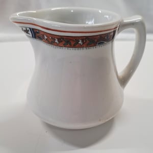 May include: White ceramic creamer with a handle and a decorative band around the rim. The band features a pattern of orange, green, and blue floral and leaf designs. The creamer has a curved body and a pouring spout.