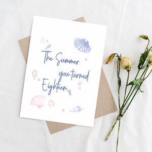 May include: A white greeting card with the text "The Summer you turned Eighteen" in blue script, decorated with watercolor seashells. The card rests on a brown envelope, with dried flowers to the side.