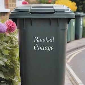 以下が含まれることがあります： ヒンジ付きの蓋が付いた濃い緑色の屋外ゴミ箱。「Bluebell Cottage」の文字が筆記体でゴミ箱の前面に書かれています。背景には他の緑色のゴミ箱と色とりどりの花が見えます。