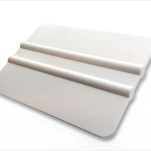 Puede incluir: Una espátula de plástico blanco con dos crestas elevadas. La herramienta tiene forma rectangular con esquinas redondeadas y un pequeño agujero cerca del borde. La espátula está diseñada para alisar superficies.