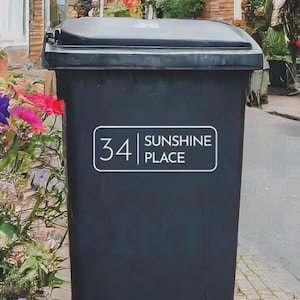 Puede incluir: Un contenedor de basura negro con tapa con bisagras. Una etiqueta rectangular blanca en la parte delantera muestra el número "34" y las palabras "SUNSHINE PLACE". El contenedor está rodeado de flores coloridas y vegetación.