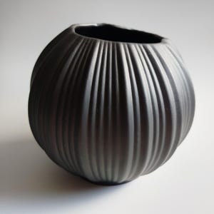 Puede incluir: Un jarrón esférico de color negro mate que presenta una serie de crestas verticales en relieve que van desde la abertura superior hasta la base. El objeto tiene un cuerpo ancho y redondeado y una abertura circular, creando una superficie acanalada.