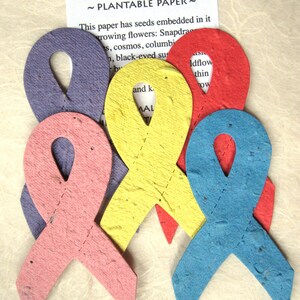 K&ouml;nnte beinhalten: Vier pflanzbare Papierbandformen in verschiedenen Farben: Lila, Rosa, Gelb und Blau. Das Papier enth&auml;lt eingebettete Samen und kann gepflanzt werden, um Blumen wachsen zu lassen. Der Text auf dem Papier lautet "Pflanzbares Papier" und listet die Arten von Blumen auf, die wachsen k&ouml;nnen.