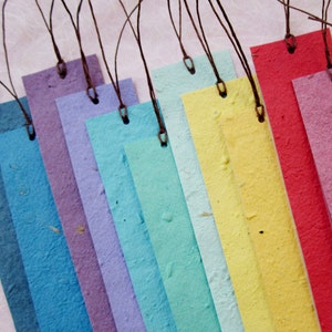 May include: A set of ten colorful bookmarks made from recycled paper with brown string attached. The bookmarks are arranged in a rainbow pattern, starting with white and ending with red.